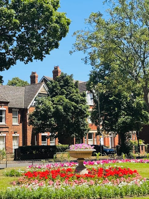 A well-maintained urban garden area in Hornchurch featuring vibrant flower beds with red, pink, and white blooms arranged around a central stone planter. The garden has neatly trimmed grass and a pave